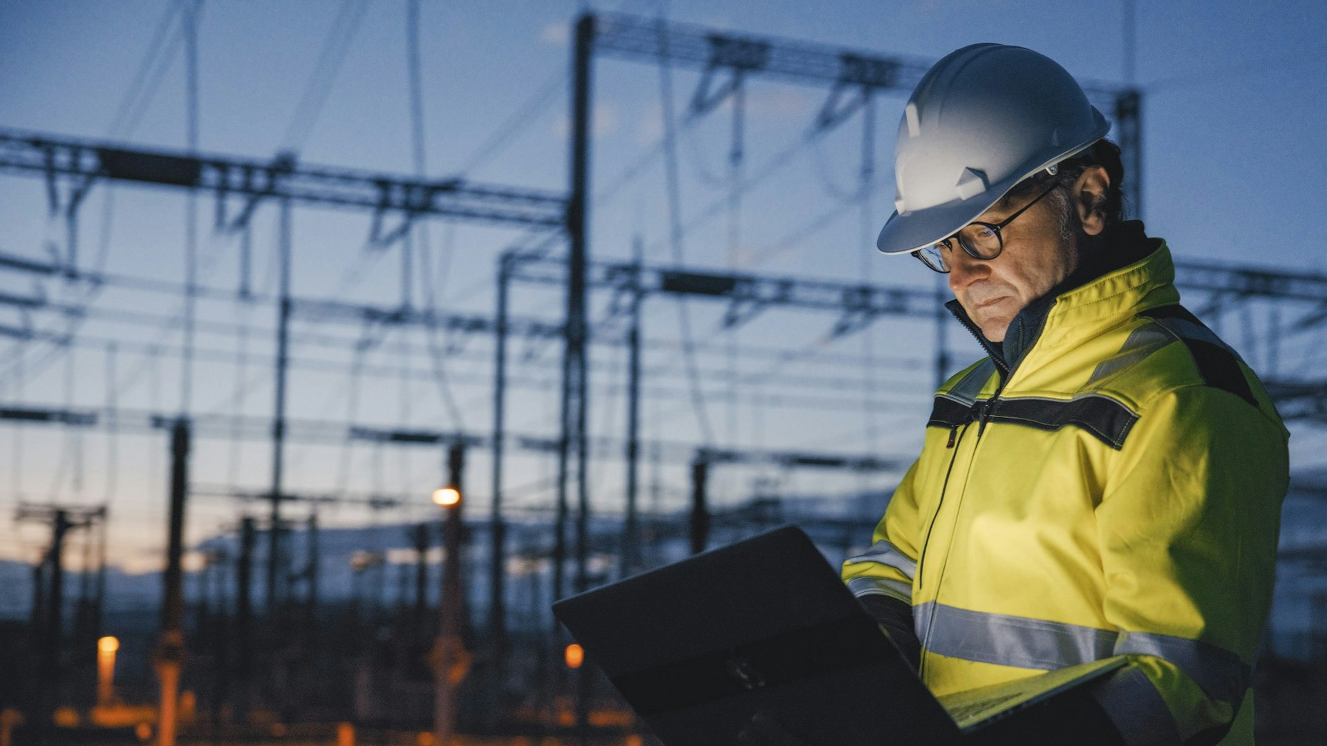 Un travailleur en tenue de sécurité se tient devant une centrale électrique, portant un casque de sécurité et un gilet de sécurité