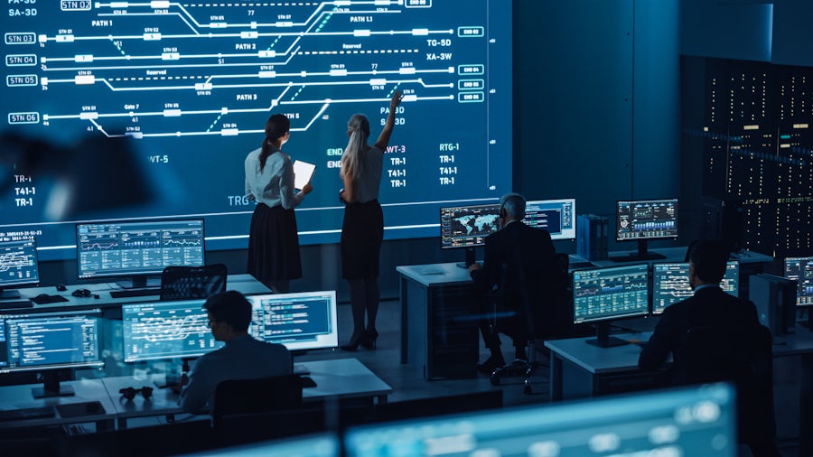 Two women look at a video wall displaying an electrical system diagram on a blue background while standing in a room with several other people working on computers.