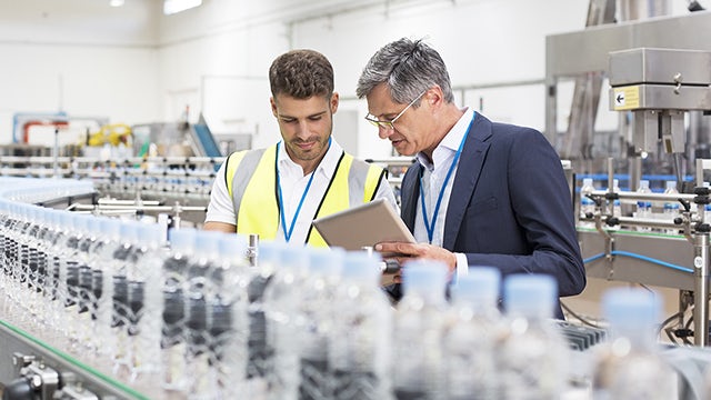Two men  with a notepad in front of a line of water bottles in a factory.