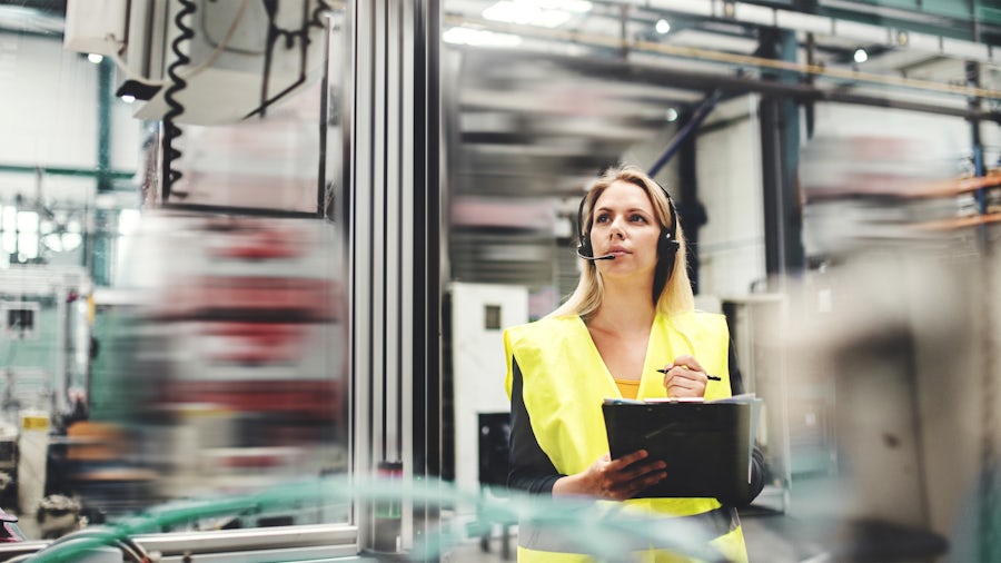 A woman with a clipboard stands on a factory floor inspecting something and prepares to write it down.