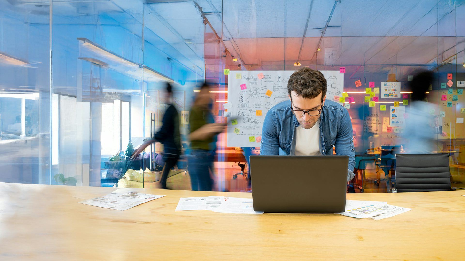 Man working on a laptop in an office, with a large whiteboard covered in sticky notes and people moving in the background.