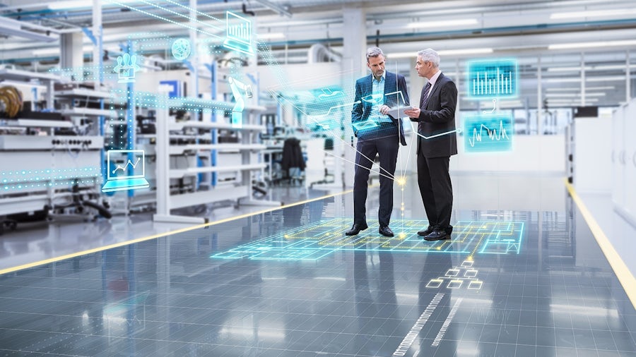 Two men in suits standing on a shop floor with a rablet looking at information. Digital graphics surround them showing data.