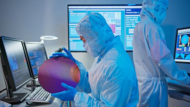 Two people wearing clean-room bunny suits. One person is holding and examining an IC wafer. The other is facing the other direction looking at a computer screen.