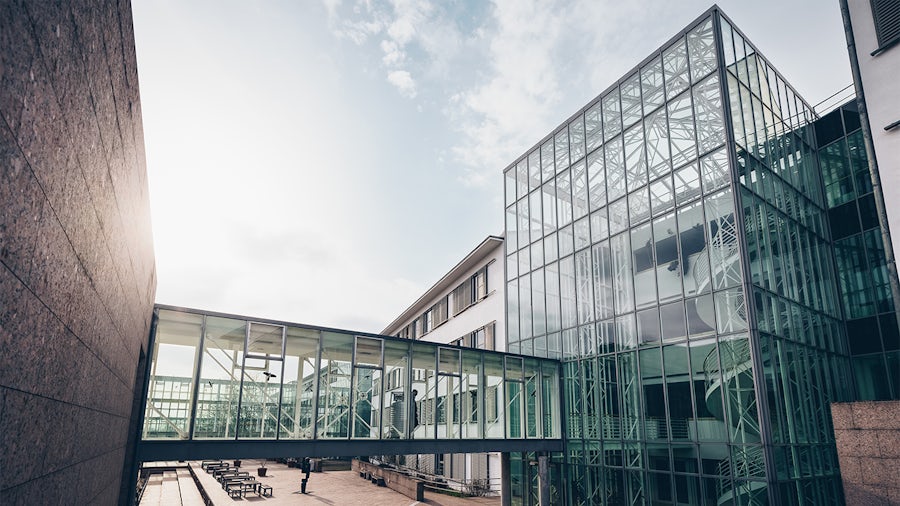 A modern architectural complex featuring a glass skywalk connecting a building with a textured dark facade to a multi-story, all-glass building. An outdoor courtyard with seating is below, under a bright, partly cloudy sky.