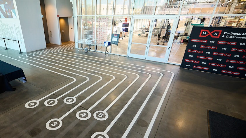 The image depicts a sleek indoor space with white floor markings guiding toward a glass-walled area. A black backdrop displays red and white logos and the text 