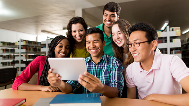 Grupa roześmianych studentów w bibliotece patrzących na tablet.