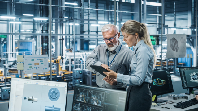 A man and a woman review data on a laptop in a room with a view of a factory floor
