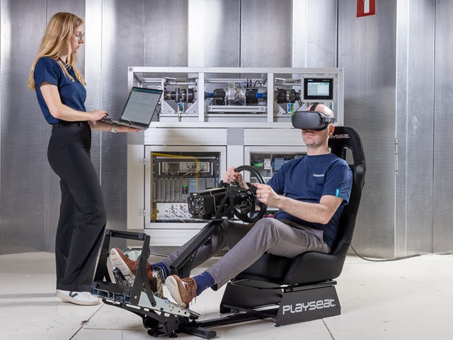 Engineers conducting vehicle testing using VR headset and playseat simulator, with one engineer monitoring data on laptop while another operates steering controls in immersive environment.