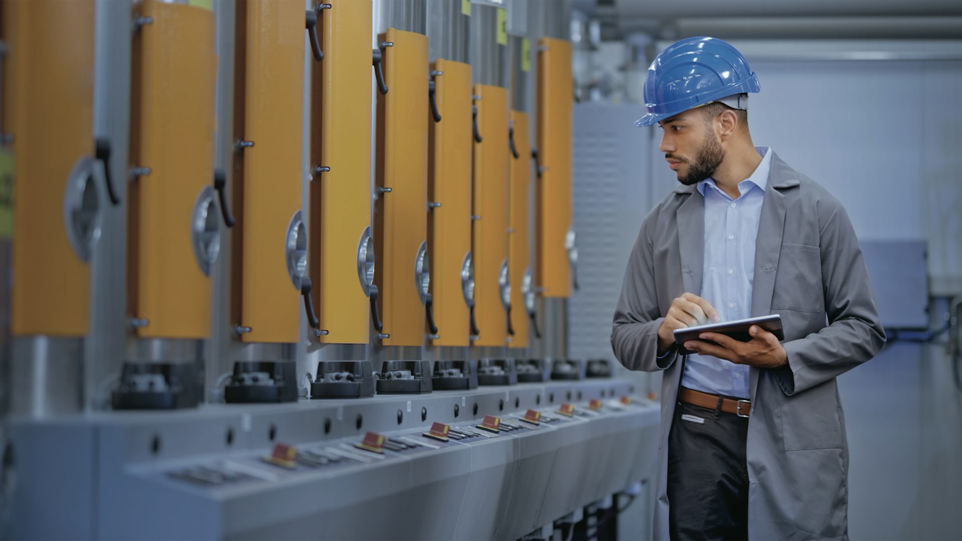 Muž v přilbě a tablet držící kontrolu výrobních strojů v průmyslovém prostředí.