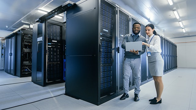 Two young professionals are standing in a server room with several rows of sever storage. One person is holding a laptop, and they are both looking at the laptop screen.