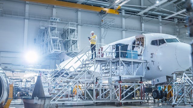 A worker stands on a platform looking at a tablet with an airplane being constructed behind him