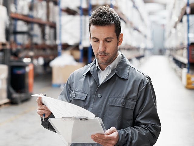 A man in a warehouse looks over papers attached to a clipboard