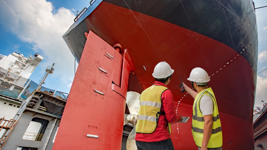 Two men in hard hats and yellow safety vests look up at the red and black hull of a large cargo ship in dry dock.