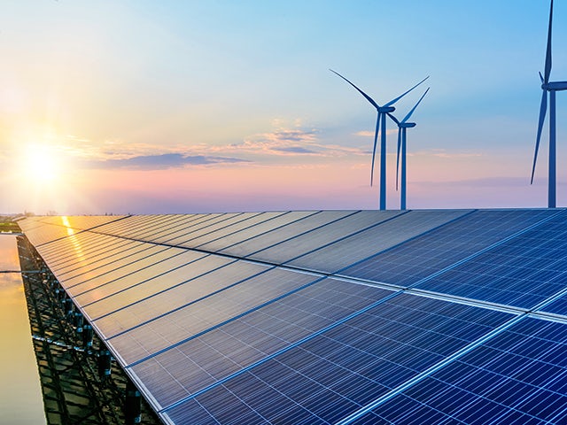 Sunset behind a row of solar panels with wind turbines in the background. 