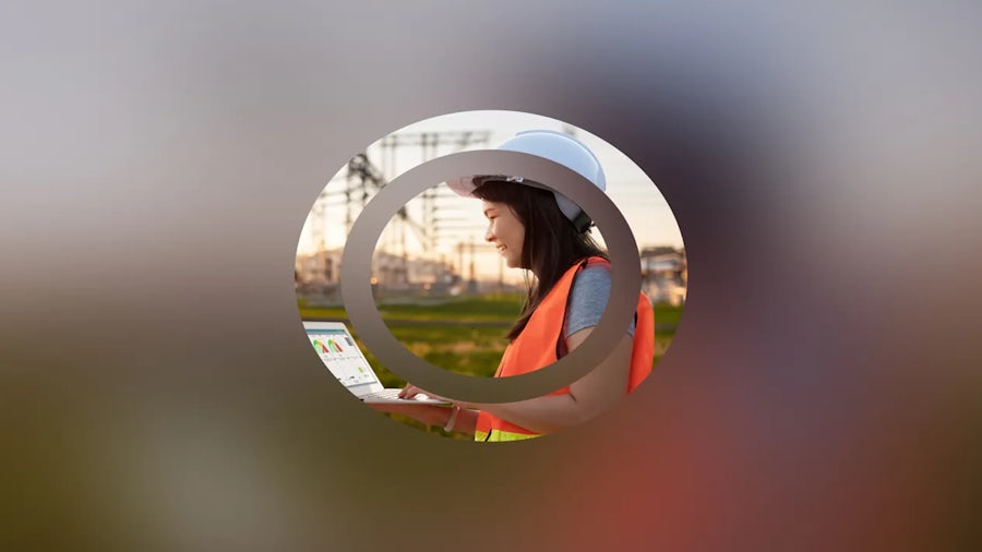 Person in a white hard hat and orange safety vest working on a laptop outdoors in an industrial area with power lines and structures in the background.