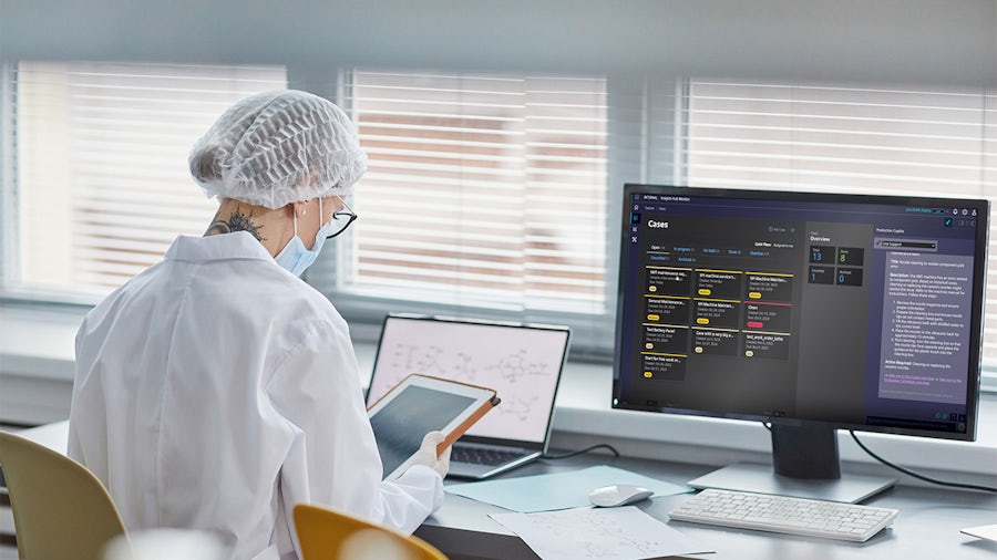 Eine Frau sitzt an einem Tisch mit Computer und hält ein Tablet in der Hand.