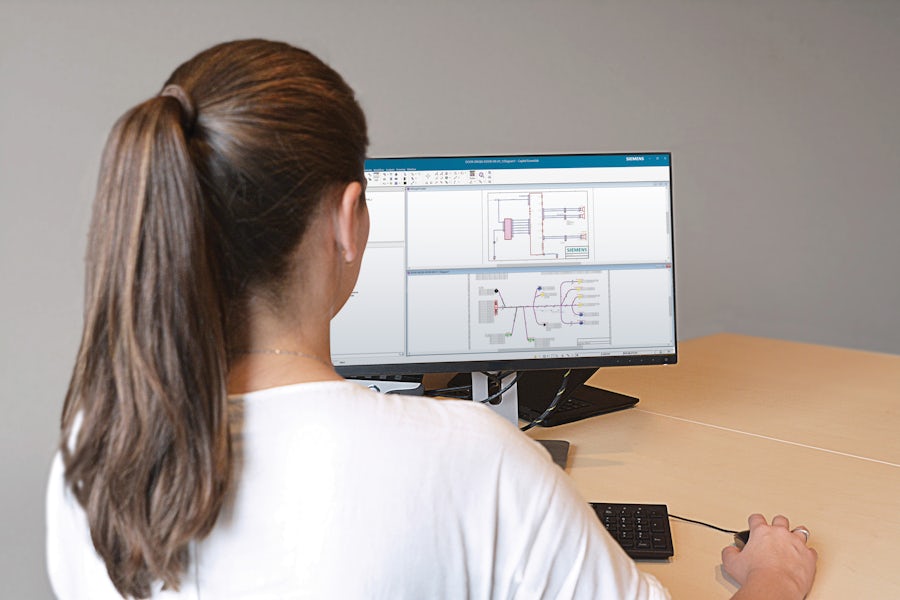 A woman sits at a desk with a computer screen displaying a wiring harness in Capital Essentials software.