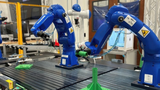 The image depicts a laboratory or industrial environment featuring two blue robotic arms working over a conveyor belt or surface. These arms are equipped with tools and sensors, interacting with several green objects, while other machinery and safety barriers are visible in the background.