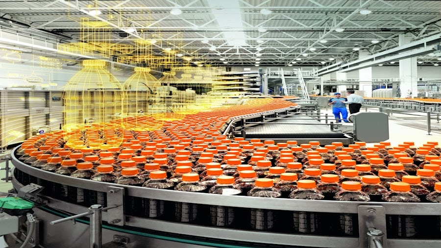 An industrial beverage bottling plant showing rows of bottles with orange caps moving on a conveyor belt, with a digital wireframe overlay representing a digital twin or smart manufacturing process. Two workers are visible in the background.