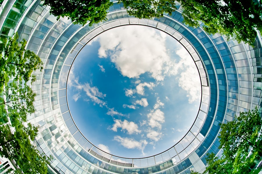 Fisheye view of a circular building with a clear blue sky in the center, surrounded by geometric glass panels and green trees.