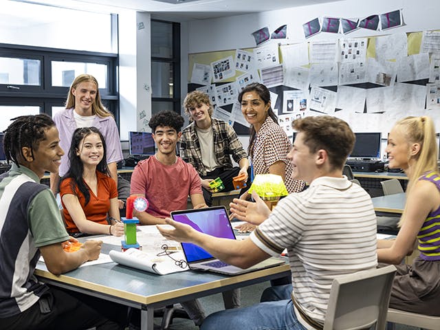 Des étudiants souriants dans une salle de classe, assis autour d’un ordinateur portable et ayant une discussion