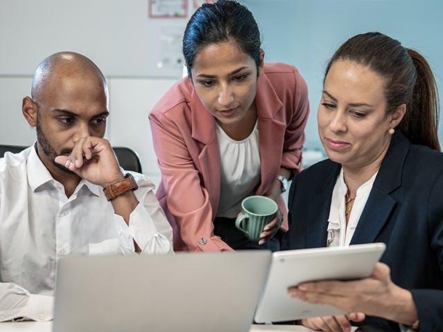 Three people in an office compare notes between a laptop and a tablet