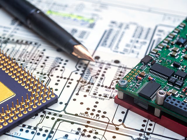 Close-up of electronic components, including a microprocessor, a printed circuit board, and a pen, placed on a detailed circuit diagram.