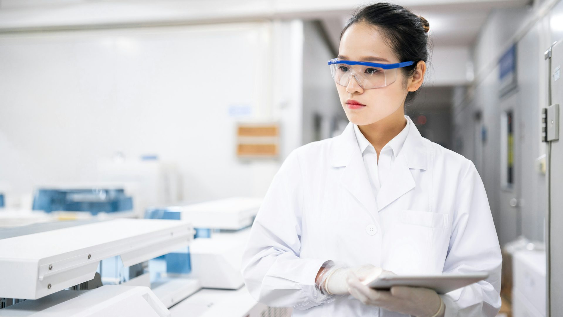 Woman wearing white lab coat and protective eye gear walking through a laboratory holding a tablet ensuring medical device compliance.