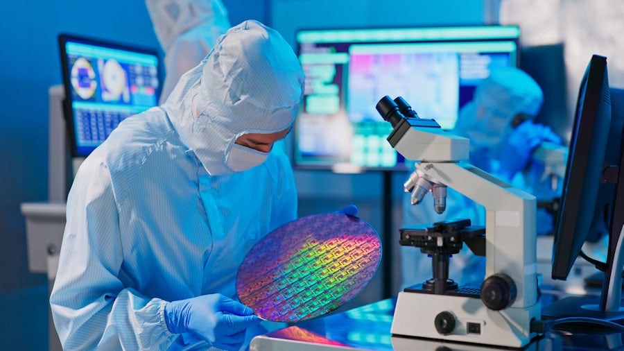 person in a  blue-tinted silicon manufacturing clean room holding a large silicon wafer that is showing a rainbow color spectrum