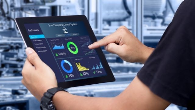 In an industrial environment, a man holds a tablet displaying graphs and data.