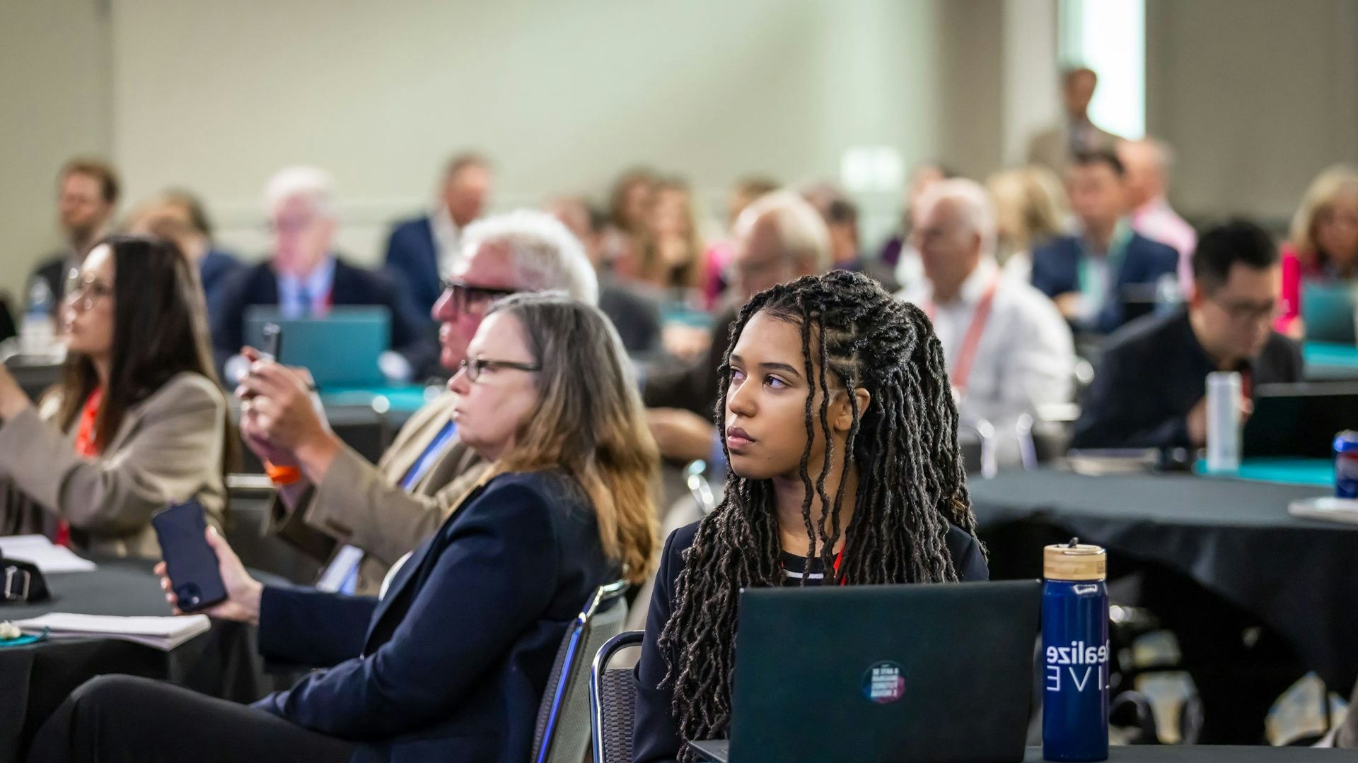 Business professionals attending corporate conference session, actively engaged with laptops and digital devices in modern meeting venue with attendees.