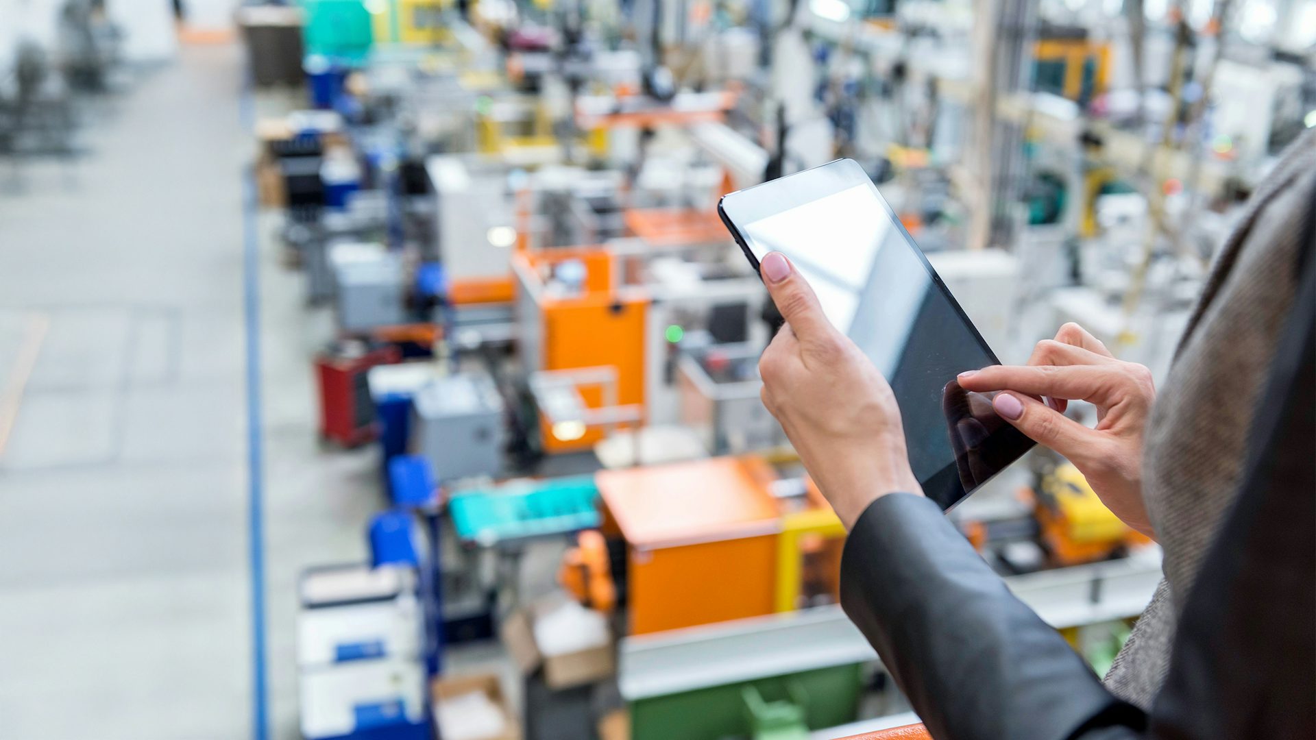 Person using a digital tablet in a modern industrial facility, with a blurred view of colorful factory machinery and equipment in the background.