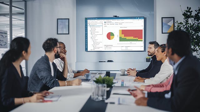 Professionals discussing quality planning using QMS software, with charts and data displayed on a screen in a meeting room.