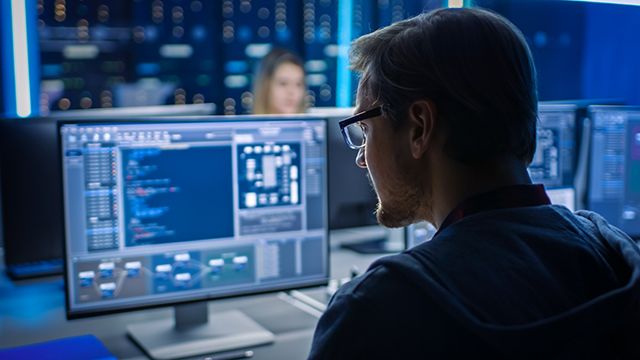 Male IT professional works on a desktop computer in a data center control room alongside a team of young programmers writing code.