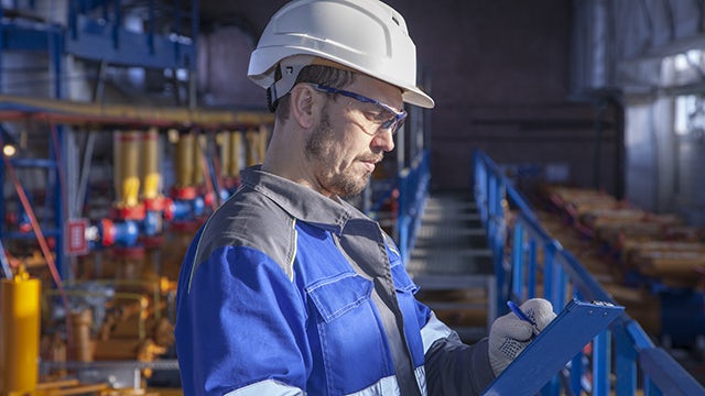 A worker wearing a blue jacket, white hardhat and safety glasses is standing in a manufacturing facility holding a blue clipboard and reviewing the content on the clipboard.