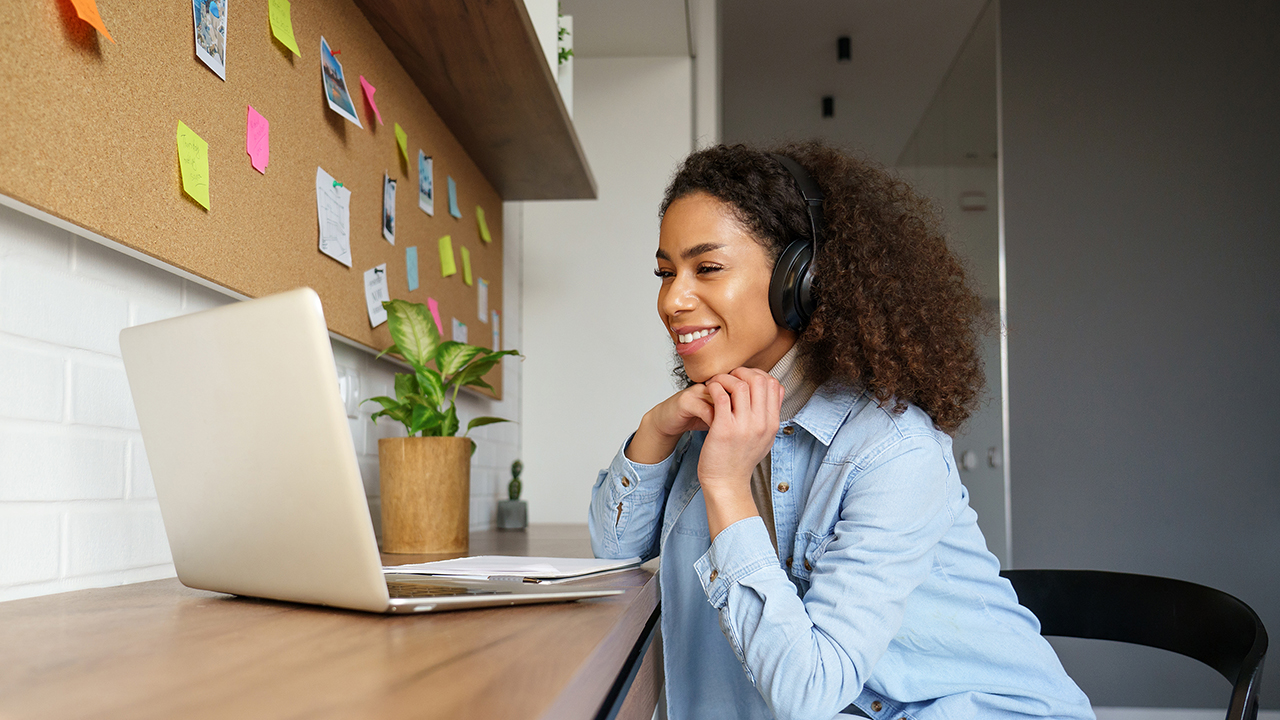 Donna con le cuffie che guarda qualcosa su un laptop. C'è una bacheca con post-it sullo sfondo.