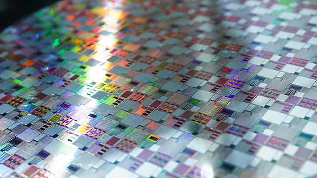 Macro photograph of silicon wafer circuitry showing detailed mosaic of integrated circuits with rainbow light diffraction creating multicolored patterns across the metallic surface.