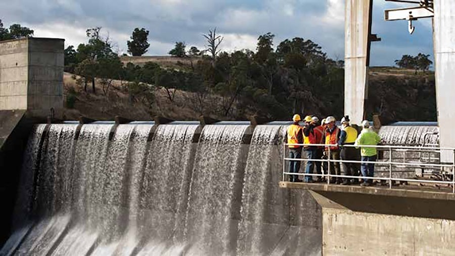 A group of engineers in hard hats and high-visibility vests stands on a platform, observing water cascading over a large dam spillway. Hills and trees are visible in the background under a cloudy sky.