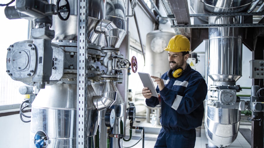 An engineer in blue coveralls and a yellow hard hat looks at a tablet. 
