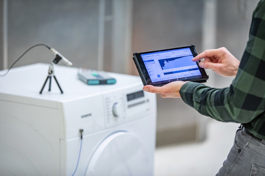 Un uomo che tiene in mano un piccolo tablet che mostra i risultati dei test di suono e vibrazione. Sullo sfondo un piccolo microfono digitale si trova su una macchina bianca in fase di test.