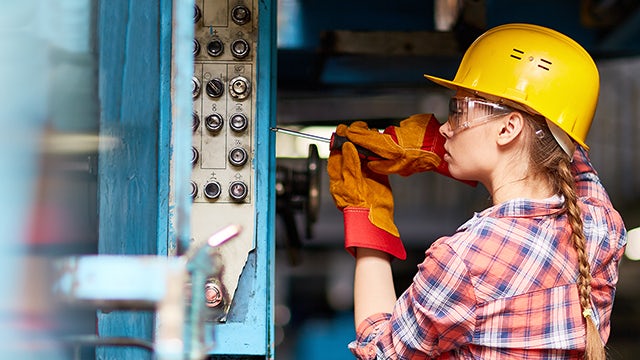 Arbeiterin mit Schutzhelm, Schutzbrille und Handschuhen bei der Arbeit in einer Fabrik